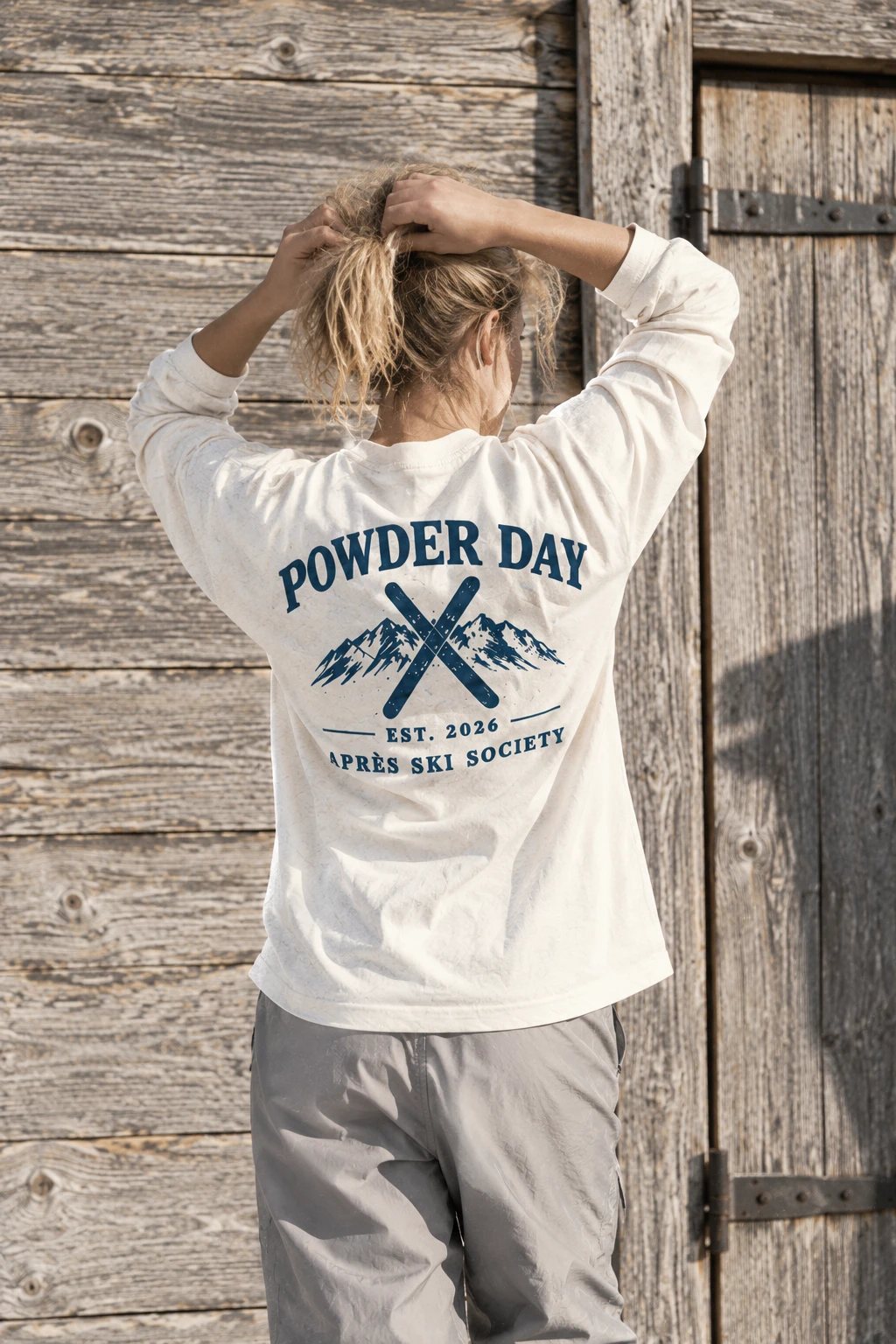 Model in de Crossed Skis longsleeve, van achteren, doet haar haar in een staart bij een houten deur