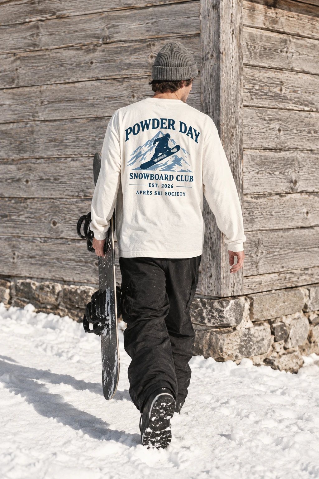 Model in de Snowboard Club longsleeve loopt met zijn snowboard langs een houten chalet