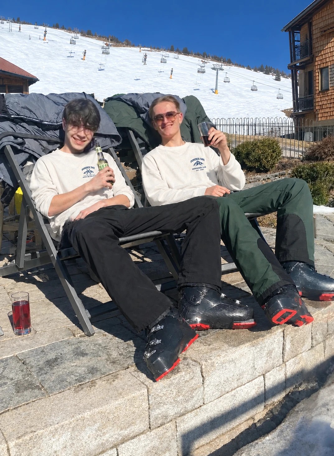 Teun en een vriend in Powder Day-longsleeves op een zonnig terras bij de piste, skischoenen nog aan
